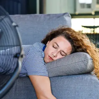 A woman lies on the couch in front of a fan, since her AC is not working.
