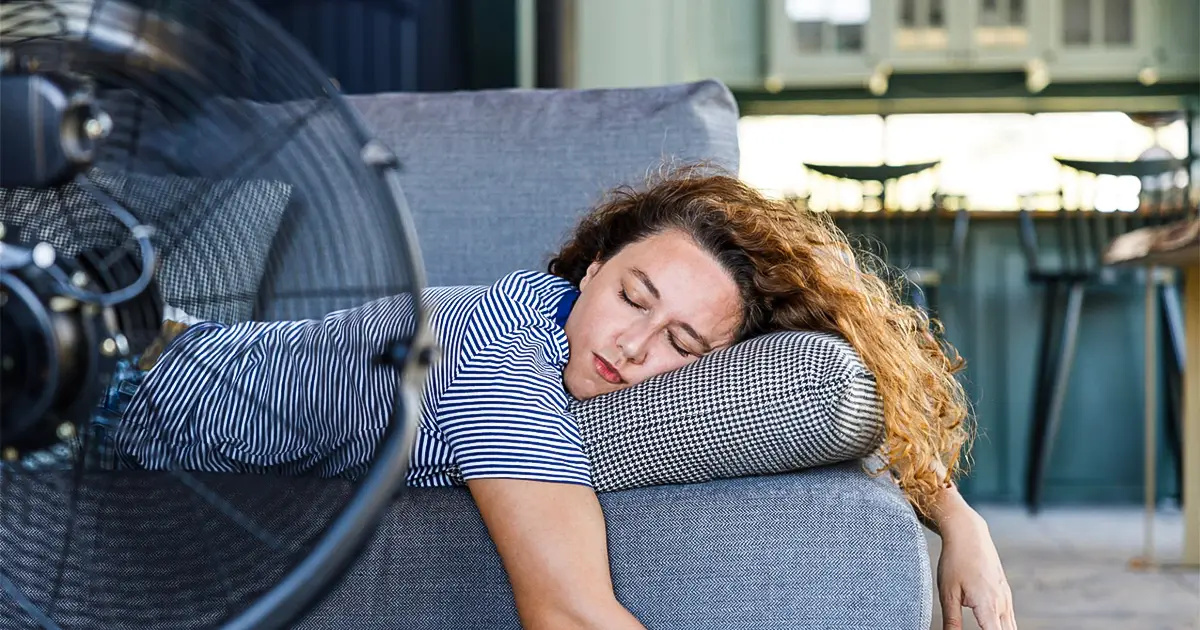 A woman lies on the couch in front of a fan, since her AC is not working.