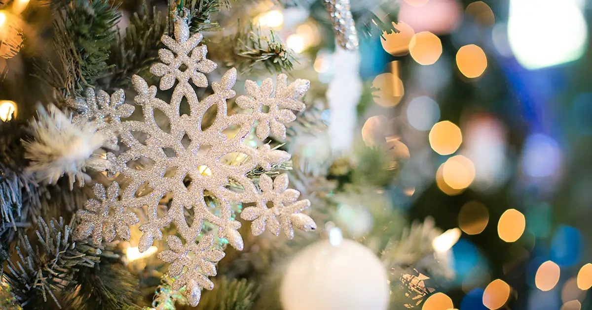 A closeup of a snowflake Christmas ornament on a tree, with dazzling lights behind.